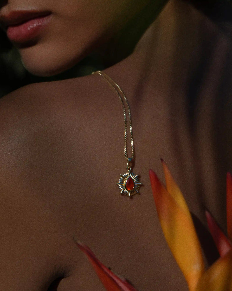 Close-up of a person wearing Awe Inspired's Aura Pendant, a gold necklace with a sun-shaped pendant radiating an orange gemstone. Orange flower petals are visible in the foreground, enhancing the aura around the wearer's chakra.