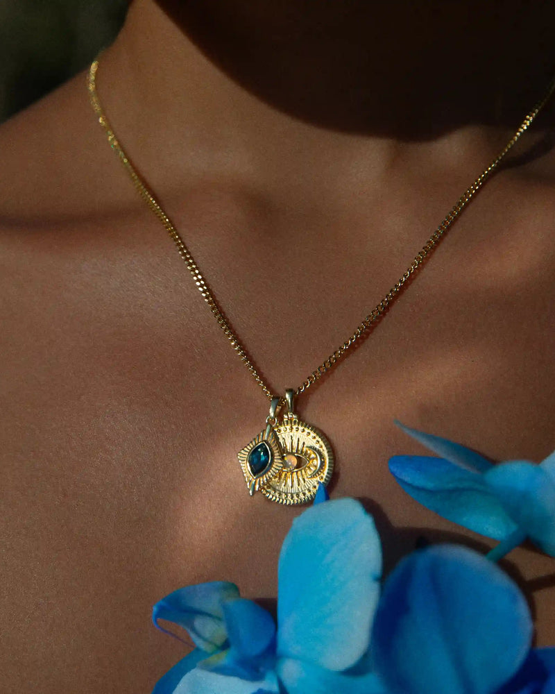 An individual's neck is adorned with the Aura Pendant from Awe Inspired, radiating an aura of elegance, while blue flowers can be seen in the foreground.