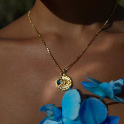 An individual's neck is adorned with the Aura Pendant from Awe Inspired, radiating an aura of elegance, while blue flowers can be seen in the foreground.