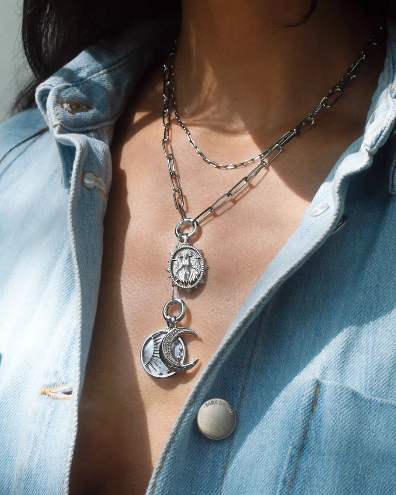 A person wearing layered silver necklaces, including the Awe Inspired Lilith Moon Charm Lariat with a moon pendant and a coin-like charm, over a partially open denim shirt.