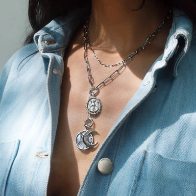 A person wearing a blue, partially buttoned shirt accessorized with the Lilith Moon Lariat Charm Necklace by Awe Inspired, featuring layered silver chains with a coin-shaped pendant and multiple charm links highlighted by a moon and star pendant.