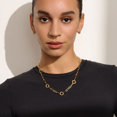 Person with a calm expression, wearing a black top, Awe Inspired's Convertible Charm Collector Necklace adorned with goddess pendants, gold earrings, and a gold septum ring, standing against a plain light-colored background.