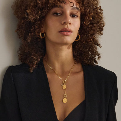 A woman with curly hair, wearing gold hoop earrings, a black outfit, and the Awe Inspired Convertible Charm Collector Necklace adorned with Goddess pendants, looks at the camera.