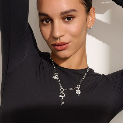 A person in a black shirt, adorned with the Awe Inspired Convertible Charm Collector Necklace featuring key and lock charms, as well as delicate goddess pendants, poses with one arm raised.