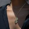 Person wearing a Cosmic Affirmation Charm Necklace by Awe Inspired, featuring an Aura pendant with two detailed geometric patterns, one with a blue stone, contrasting against a dark top.