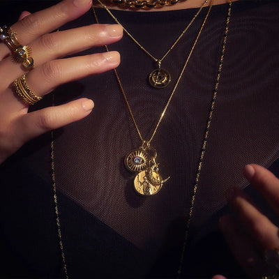 Close-up of a person wearing layered gold necklaces and rings on their fingers against a black mesh top background. Among the jewelry, an Awe Inspired Dancing in the Moonlight Charm Necklace shines prominently, accompanied by various designs of pendants and rings, including some featuring moonstone inlay.