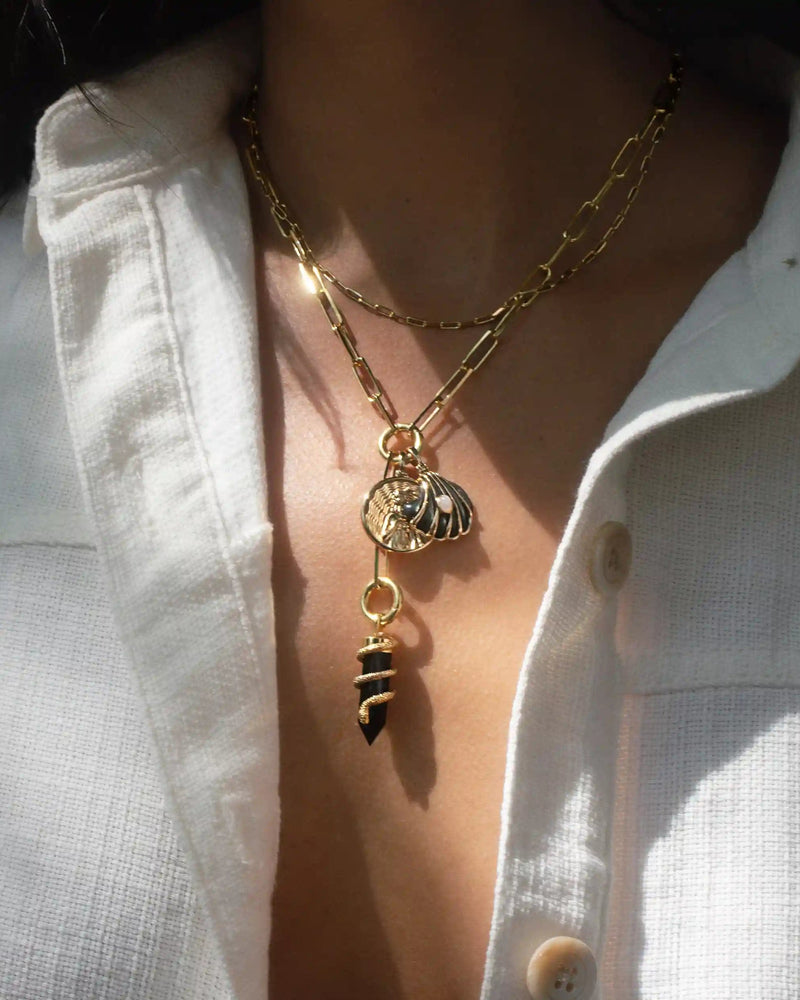 A person wearing a white shirt is adorned with the Awe Inspired Dark Waves Charm Lariat, layered gold necklaces that feature multiple charm links, including a seashell and a black and gold striped charm.