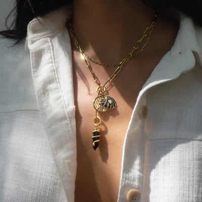 A person wearing a white shirt is adorned with the Awe Inspired Dark Waves Charm Lariat, layered gold necklaces that feature multiple charm links, including a seashell and a black and gold striped charm.