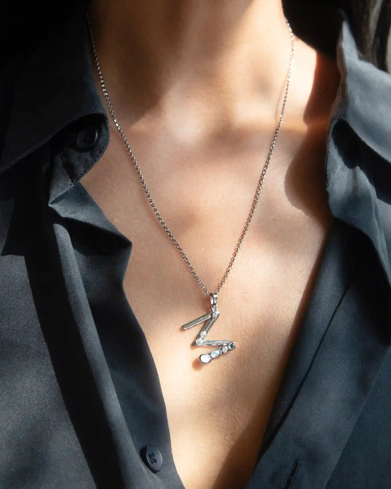 A person wearing a black shirt showcases the Awe Inspired Silver Gemstone Initial Necklace, featuring a statement size zigzag pendant adorned with bezel set gemstones.