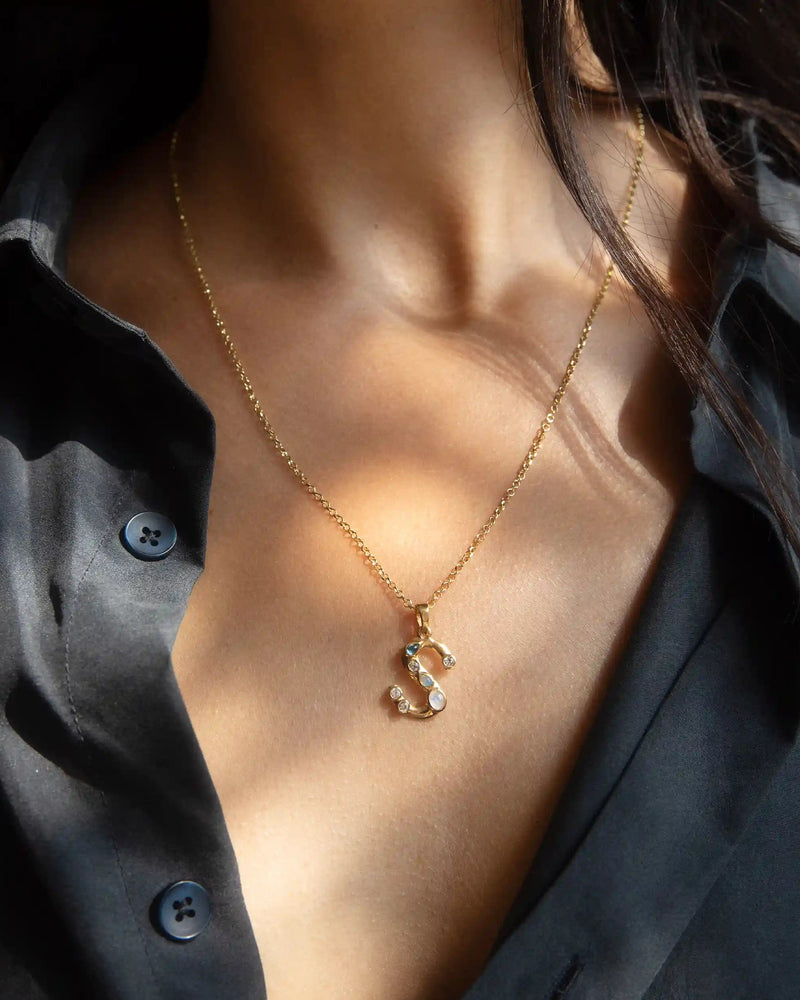 Close-up of a person wearing a black button-down shirt and the Gold Gemstone Initial Necklace by Awe Inspired, featuring a bezel-set gemstone pendant shaped like the letter 'S', adhering to statement size initial charms.