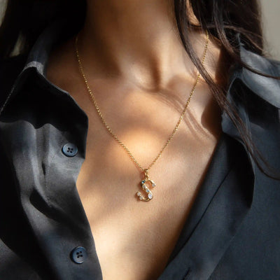 Close-up of a person wearing a black button-down shirt and the Gold Gemstone Initial Necklace by Awe Inspired, featuring a bezel-set gemstone pendant shaped like the letter 'S', adhering to statement size initial charms.