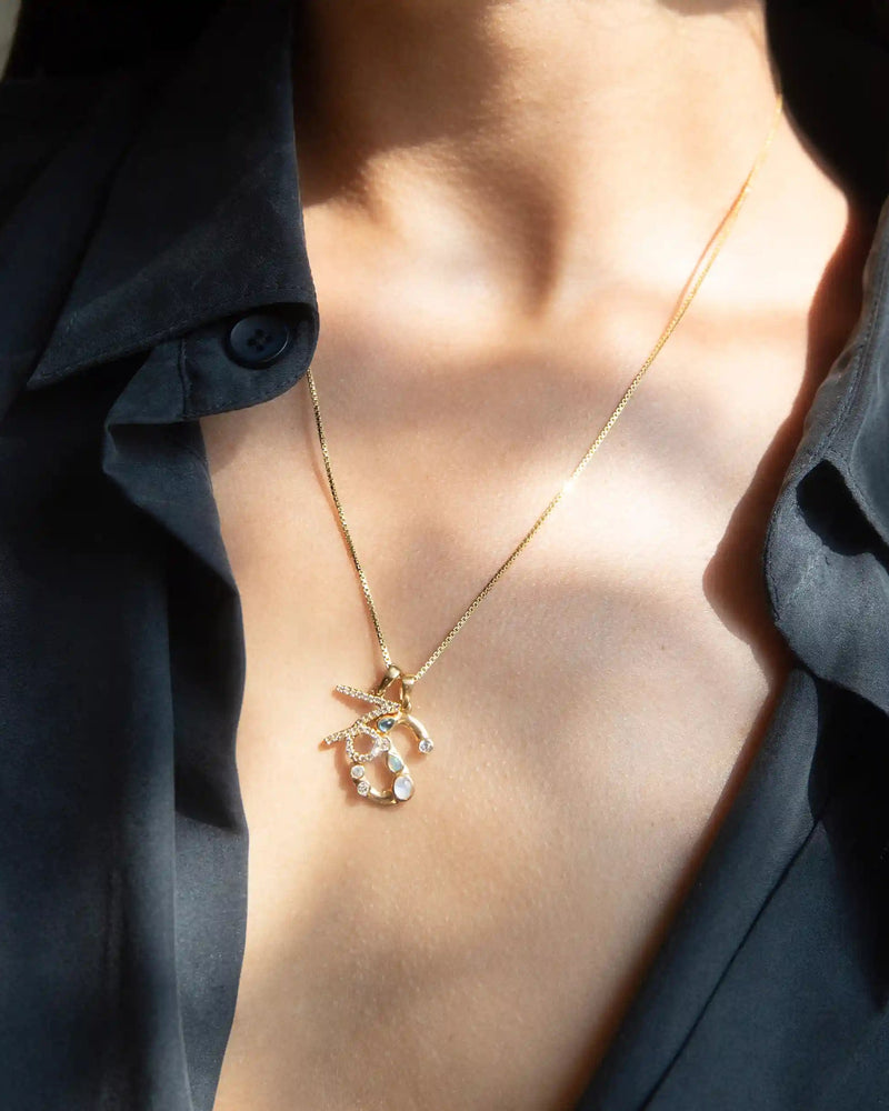 A person wearing a dark, partially open shirt and the Awe Inspired Gold Gemstone Initial Necklace, which boasts statement size initial charms alongside a pendant featuring starfish and shell designs.