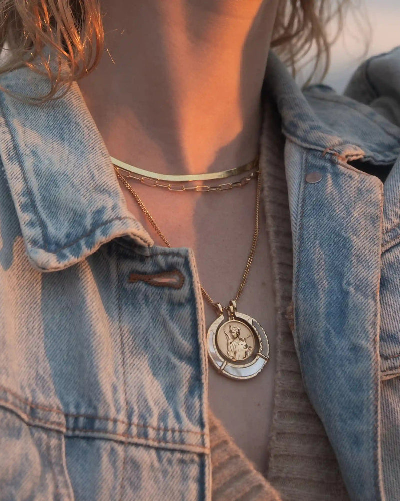 Close-up of a person wearing a denim jacket and layered necklaces, featuring the Hunter's Moon Halo Necklace Set from Awe Inspired, which includes a gold choker and an Artemis pendant with a circular design believed to absorb negative energy.