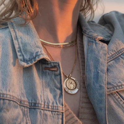 Close-up of a person wearing a denim jacket and layered necklaces, featuring the Hunter's Moon Halo Necklace Set from Awe Inspired, which includes a gold choker and an Artemis pendant with a circular design believed to absorb negative energy.