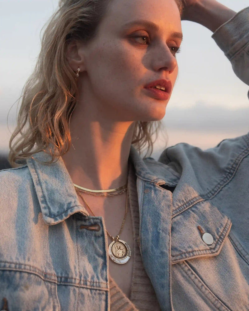 A person with wavy hair wearing a denim jacket and layered necklaces, including the Hunter's Moon Halo Necklace Set from Awe Inspired, gazes into the distance with one hand touching their head.