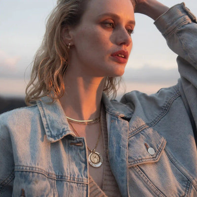 A person with wavy hair wearing a denim jacket and layered necklaces, including the Hunter's Moon Halo Necklace Set from Awe Inspired, gazes into the distance with one hand touching their head.