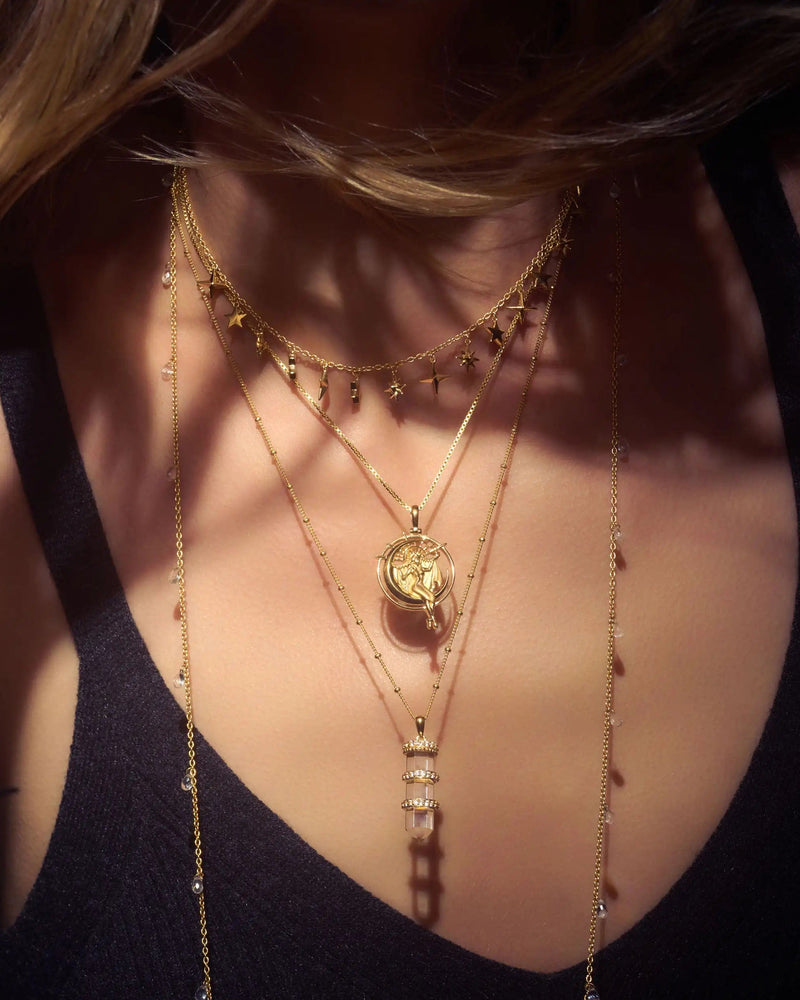 A person wearing multiple layered gold necklaces, including ones with small charms and an Awe Inspired Manifestation Crystal Necklace featuring a clear quartz pendant, on a black top.