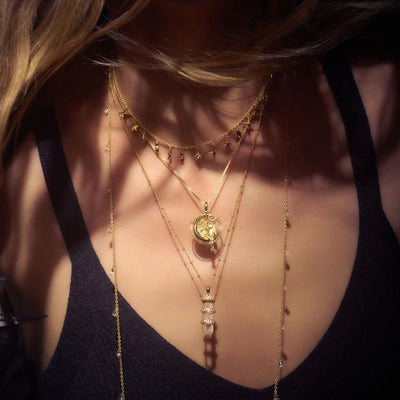 A person wearing multiple layered gold necklaces, including ones with small charms and an Awe Inspired Manifestation Crystal Necklace featuring a clear quartz pendant, on a black top.