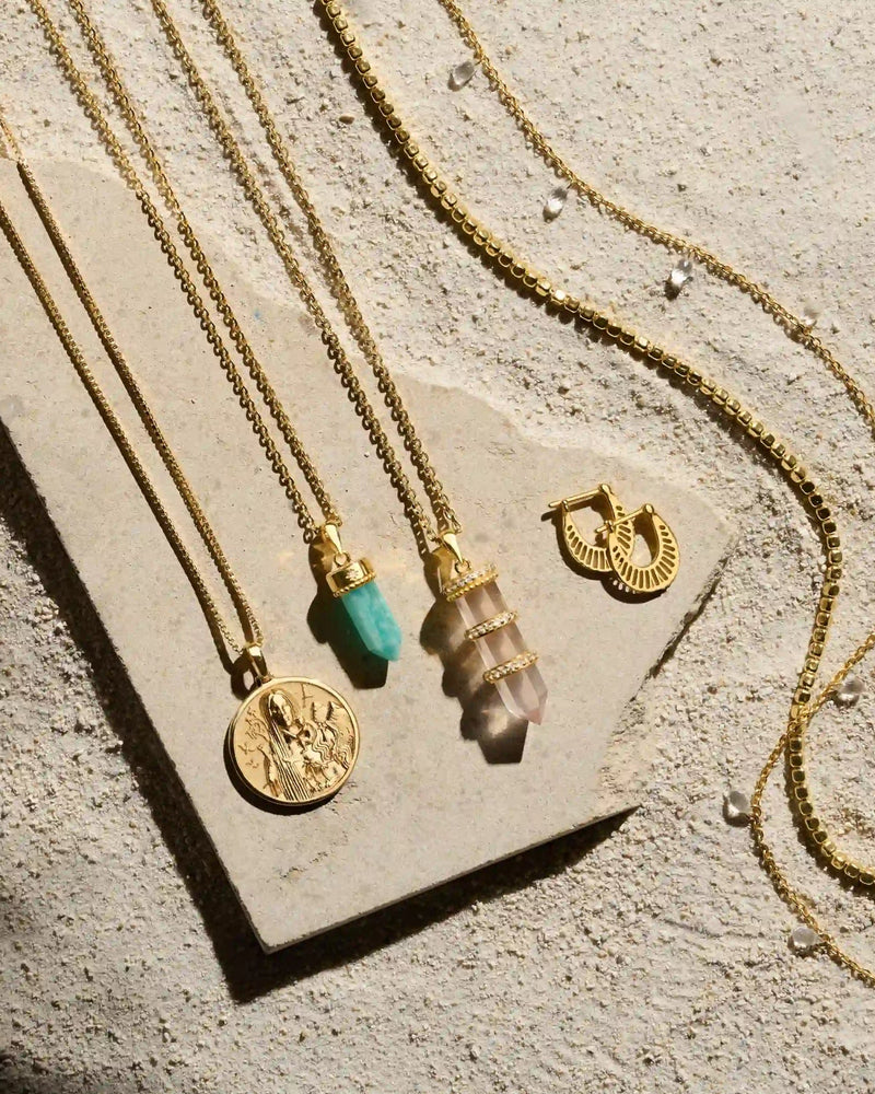 Four gold necklaces and a pair of gold hoop earrings are displayed on a textured stone surface. The necklaces, including the Manifestation Crystal Necklace by Awe Inspired, feature various pendants such as a turquoise stone, a layered crystal, and a protective clear quartz.
