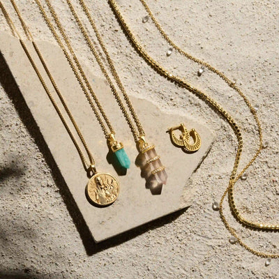 Four gold necklaces and a pair of gold hoop earrings are displayed on a textured stone surface. The necklaces, including the Manifestation Crystal Necklace by Awe Inspired, feature various pendants such as a turquoise stone, a layered crystal, and a protective clear quartz.