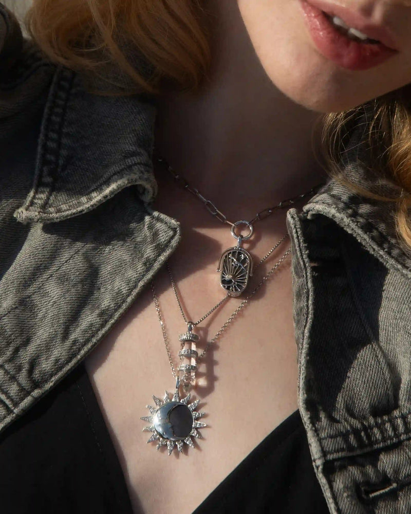 Close-up of a person wearing a denim jacket, a black top, and layered silver necklaces featuring a sun pendant, intricate designs, and Awe Inspired's Manifestation Crystal Necklace with clear quartz.