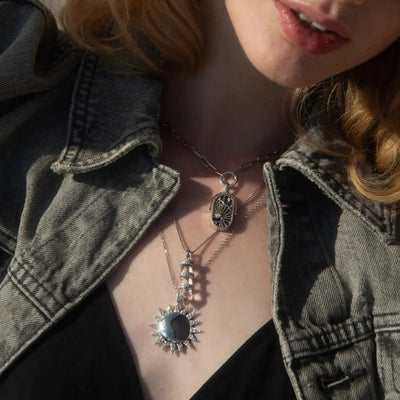 Close-up of a person wearing a denim jacket, a black top, and layered silver necklaces featuring a sun pendant, intricate designs, and Awe Inspired's Manifestation Crystal Necklace with clear quartz.