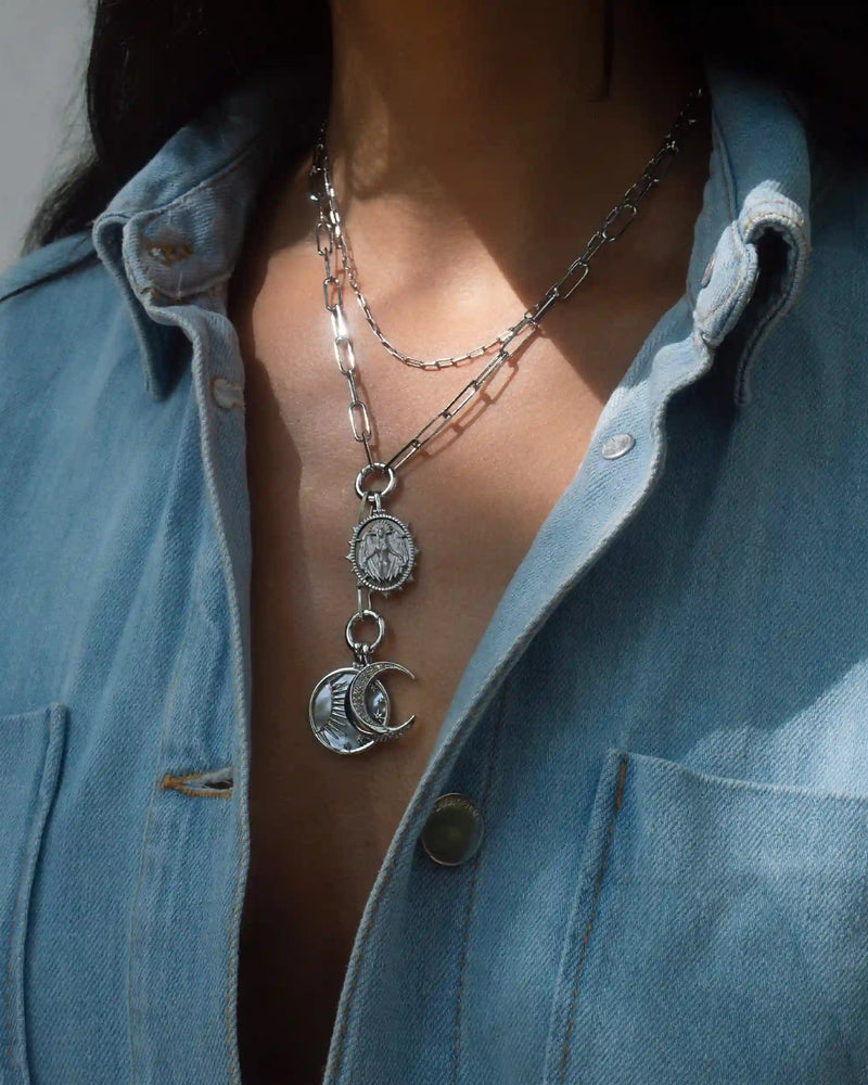 Close-up of a person wearing a denim shirt and two layered silver chain necklaces, one with a circular pendant and the other with the Awe Inspired Lilith Moon Charm Lariat featuring a crescent moon charm.