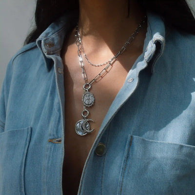 Close-up of a person wearing a denim shirt and layered silver necklaces, including the Lilith Moon Lariat Charm Necklace by Awe Inspired, featuring a crescent moon and coin pendant.