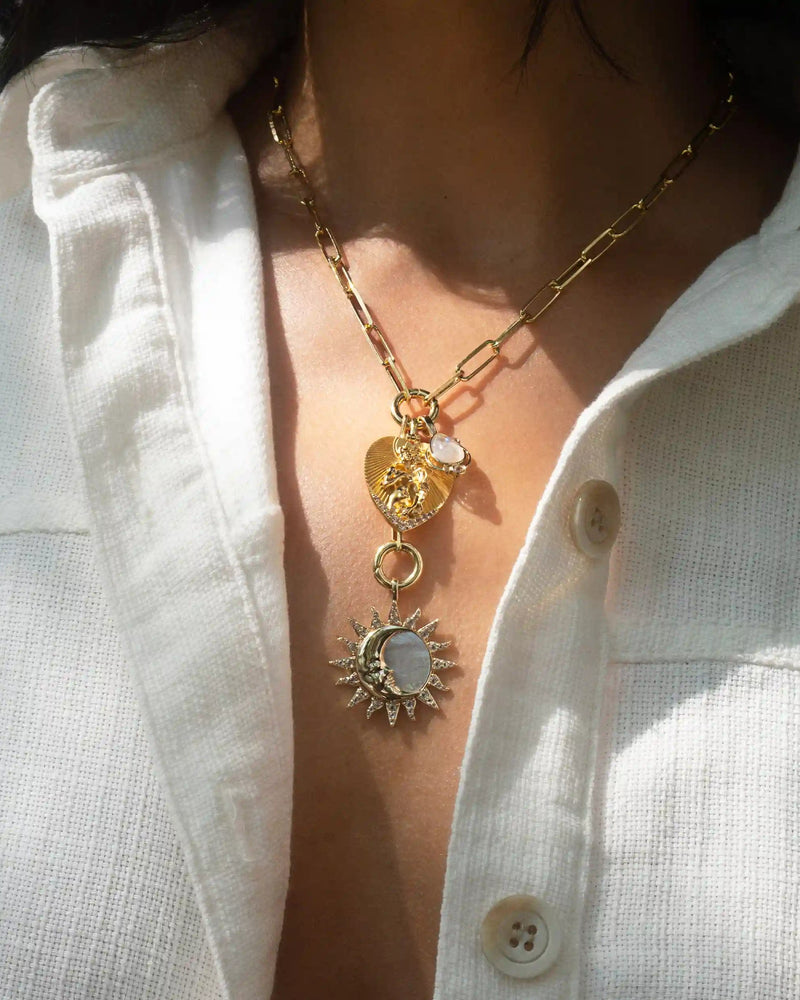 A person wearing a white button-up shirt is displaying the Love Goddess Charm Lariat by Awe Inspired, featuring layered gold necklaces adorned with celestial motifs, including heart and sun pendants.