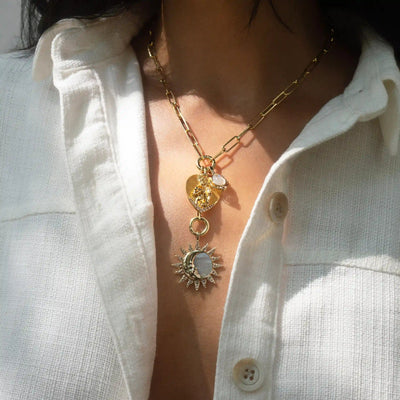 A person wearing a white button-up shirt is displaying the Love Goddess Charm Lariat by Awe Inspired, featuring layered gold necklaces adorned with celestial motifs, including heart and sun pendants.