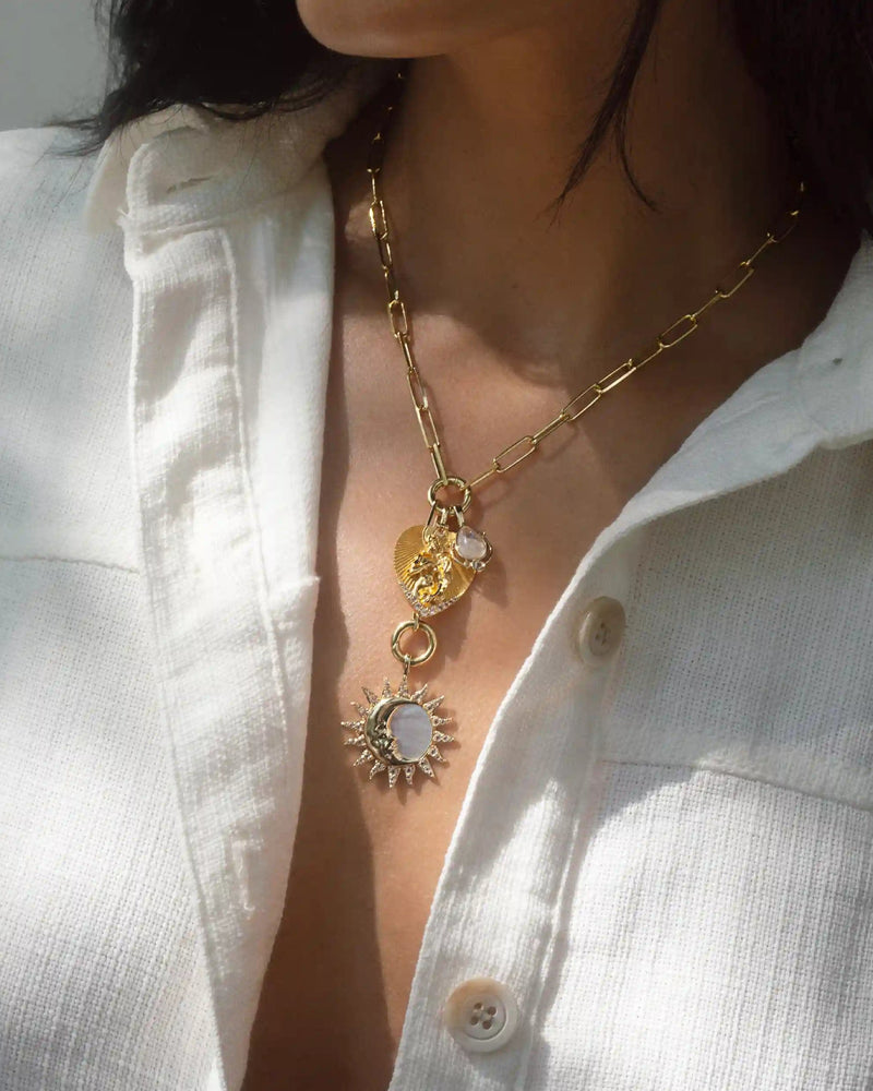 Close-up of a person wearing a white button-up shirt, showcasing two gold necklaces with celestial motifs, one featuring sun-themed pendants and the other, the Love Goddess Charm Lariat necklace by Awe Inspired with delicate sun and moon charms.