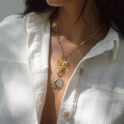 Close-up of a person wearing a white button-up shirt, showcasing two gold necklaces with celestial motifs, one featuring sun-themed pendants and the other, the Love Goddess Charm Lariat necklace by Awe Inspired with delicate sun and moon charms.