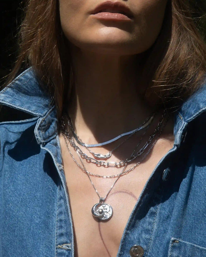 A close-up of a person wearing an Awe Inspired Statement Chain Necklace, styled as part of a chic necklace stack with pendant and chunky chain pieces, layered over a partially unbuttoned denim shirt.