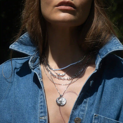 A close-up of a person wearing an Awe Inspired Statement Chain Necklace, styled as part of a chic necklace stack with pendant and chunky chain pieces, layered over a partially unbuttoned denim shirt.