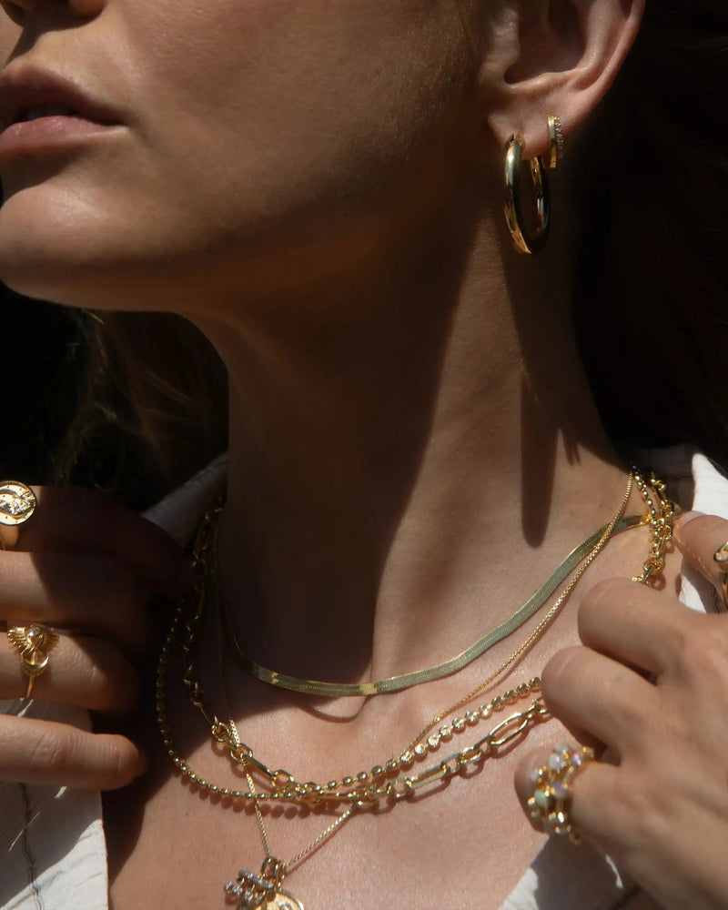 Close-up of a person wearing various gold jewelry, including hoop earrings, multiple rings, and an elegant necklace stack featuring the Statement Chain Necklace by Awe Inspired. The person is adjusting the necklaces with their hands.