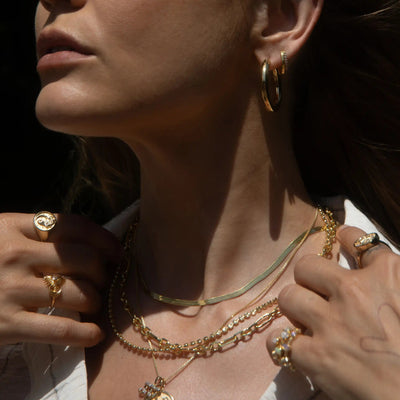 Close-up of a person wearing various gold jewelry, including hoop earrings, multiple rings, and an elegant necklace stack featuring the Statement Chain Necklace by Awe Inspired. The person is adjusting the necklaces with their hands.