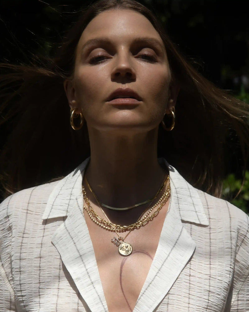 A person with long brown hair wears gold hoop earrings and the Awe Inspired Statement Chain Necklace, standing outdoors in sunlight, with eyes half-closed, wearing a white textured top.