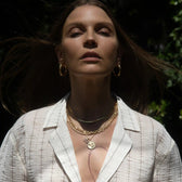 A person with long brown hair wears gold hoop earrings and the Awe Inspired Statement Chain Necklace, standing outdoors in sunlight, with eyes half-closed, wearing a white textured top.