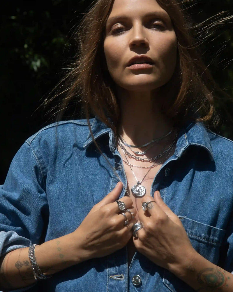 A person with long hair stands outdoors wearing a denim jacket, showcasing the Statement Chain Necklace by Awe Inspired and various rings, while holding their jacket collar with both hands.