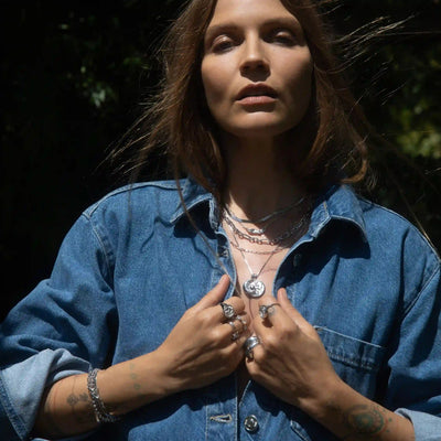 A person with long hair stands outdoors wearing a denim jacket, showcasing the Statement Chain Necklace by Awe Inspired and various rings, while holding their jacket collar with both hands.