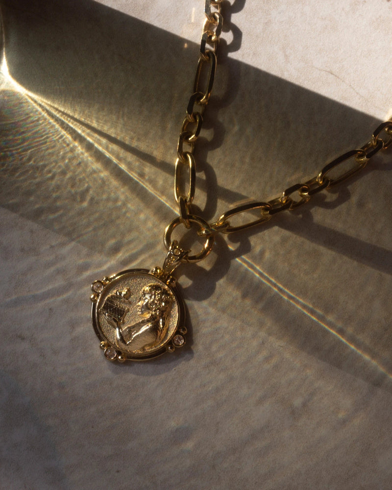 The Statement Single Charm Collector Necklace by Awe Inspired, featuring a wide luxe link chain and a round pendant with an engraved figure, is placed on a surface with shadows and light reflections.