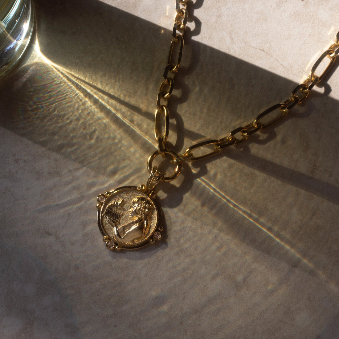 The Statement Single Charm Collector Necklace by Awe Inspired, featuring a wide luxe link chain and a round pendant with an engraved figure, is placed on a surface with shadows and light reflections.