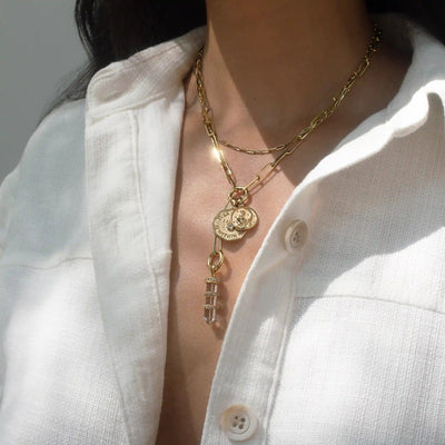 A person wearing a white textured shirt with a Manifestation Charm Lariat necklace from Awe Inspired, adorned with gold chains featuring Everything I Need Is Within Me coin pendants and a hanging cylindrical piece.