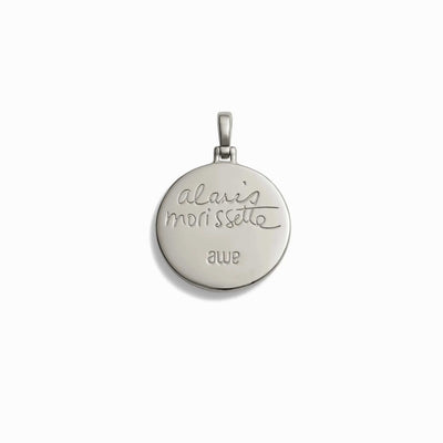 The Storm Before the Calm Charm Necklace by Awe Inspired is a round metal pendant engraved with "Alanis Morissette" in cursive and the word "awe" below it, embodying the essence of the divine feminine.