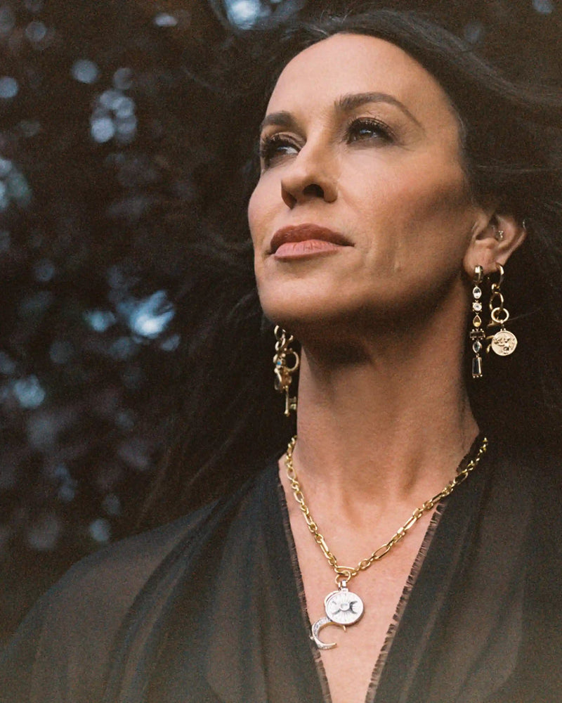 A woman with long dark hair, wearing a black top and gold jewelry that includes The Storm Before the Calm Charm Necklace by Awe Inspired, looks confidently upwards, embodying the essence of the divine feminine.