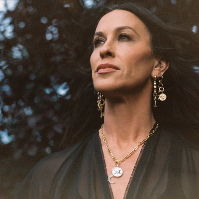 A woman with long dark hair, wearing a black top and gold jewelry that includes The Storm Before the Calm Charm Necklace by Awe Inspired, looks confidently upwards, embodying the essence of the divine feminine.