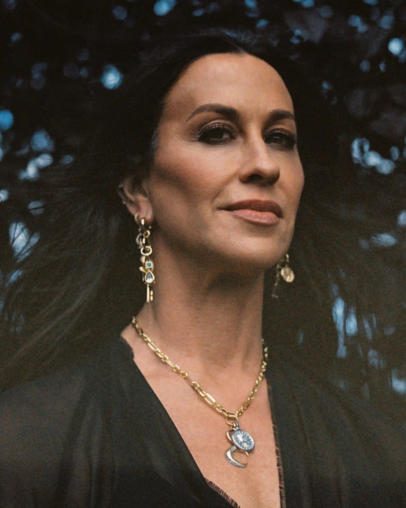 A person with long dark hair, wearing a black top and gold jewelry, including The Storm Before the Calm Charm Necklace by Awe Inspired and dangling earrings, stands against a dark background.