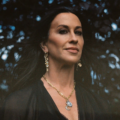 A person with long dark hair, wearing a black top and gold jewelry, including The Storm Before the Calm Charm Necklace by Awe Inspired and dangling earrings, stands against a dark background.