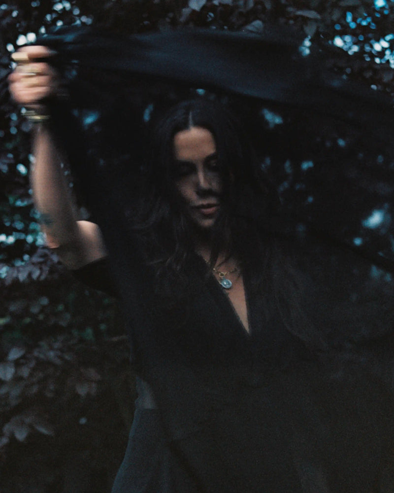 A person holding and moving a dark, translucent fabric in front of their face, surrounded by dimly lit foliage, wears The Storm Before the Calm Charm Necklace from Awe Inspired, which reflects the light with an air of divine feminine energy.