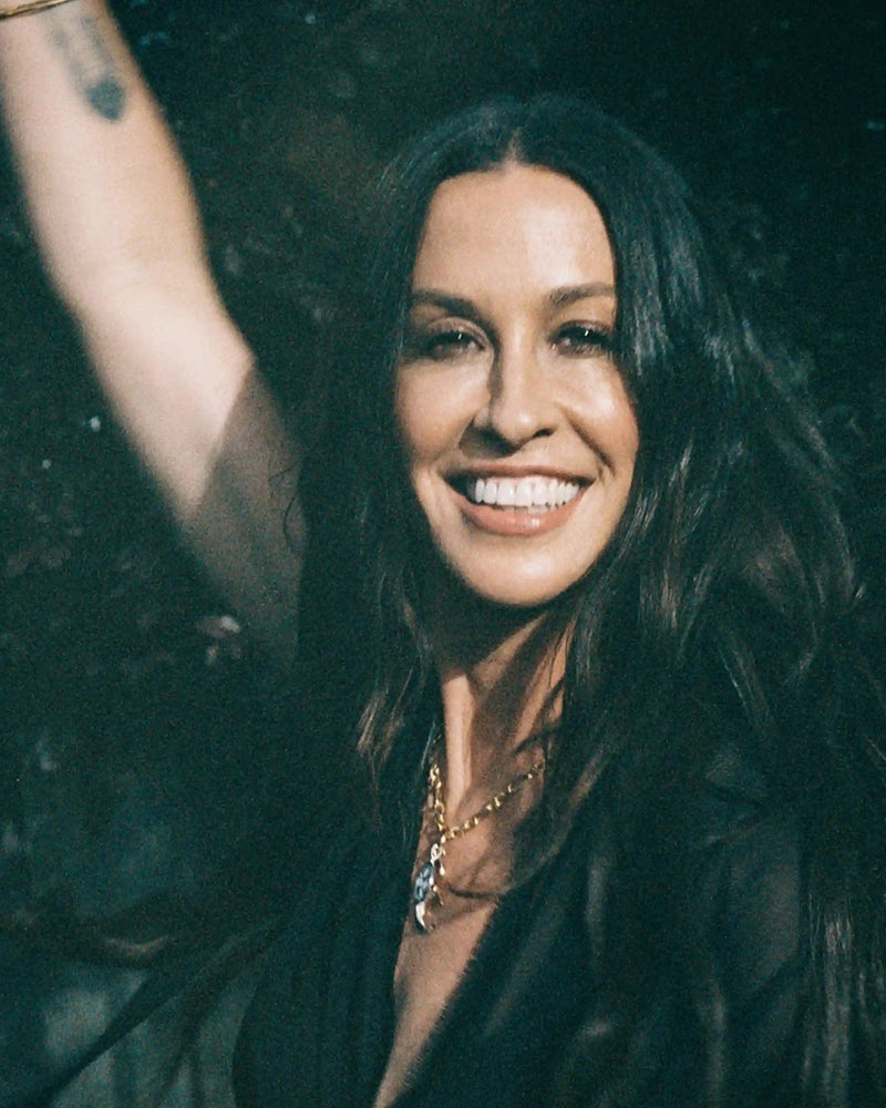 A person with long dark hair, wearing Awe Inspired's Storm Before the Calm Charm Necklace, smiles and raises one arm. The background is dark and leafy, adding to the divine feminine aura she embodies.